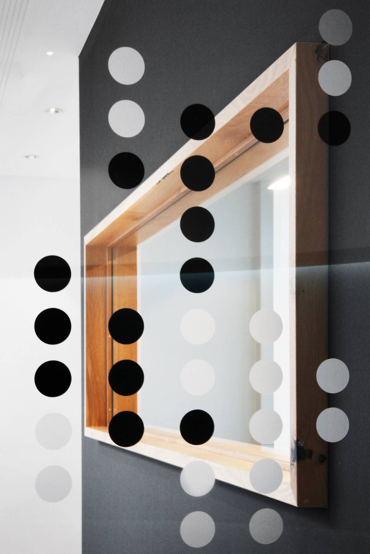 Glasscheibe mit schwarzen und weißen Punkten, dahinter ein rechteckiger, hölzerner Fensterrahmen vor einer grauen Wand.