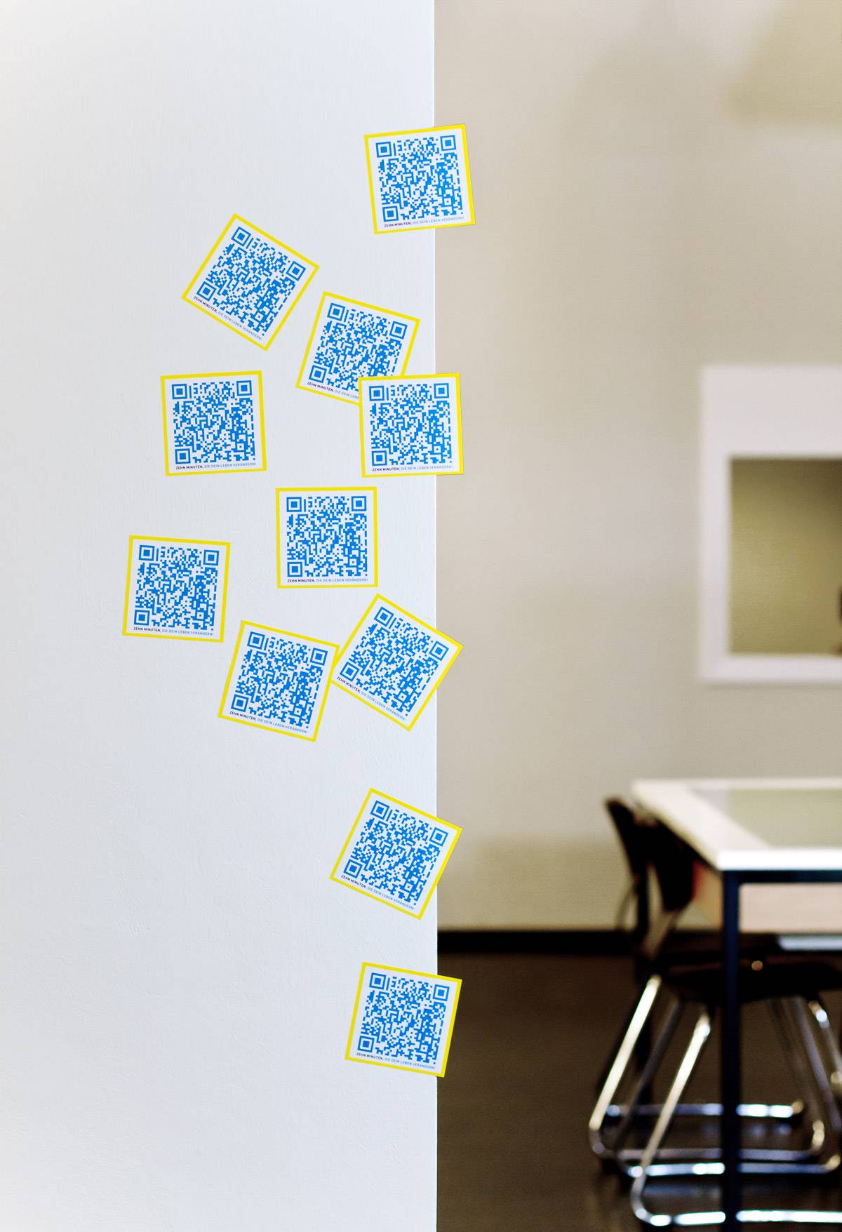 Mehrere blaue QR-Code-Aufkleber mit gelbem Rand kleben an einer weißen Wand, im Hintergrund ein Besprechungstisch mit Stühlen.