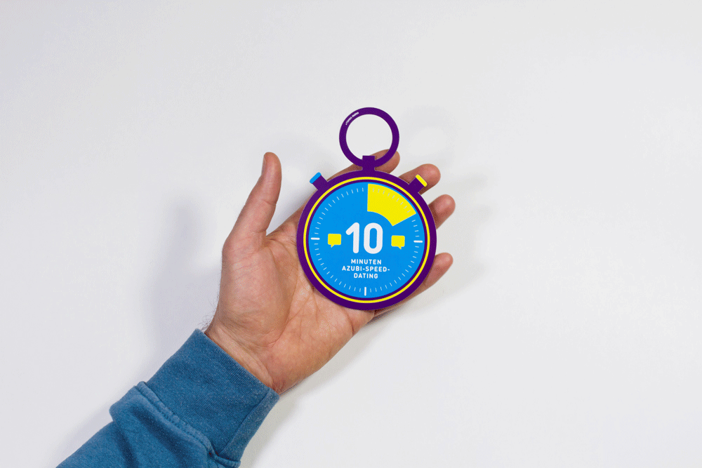 Eine Hand hält eine bunte Stoppuhr mit der Aufschrift „10 Minuten Azubi-Speed-Dating“ auf blauem Hintergrund.
