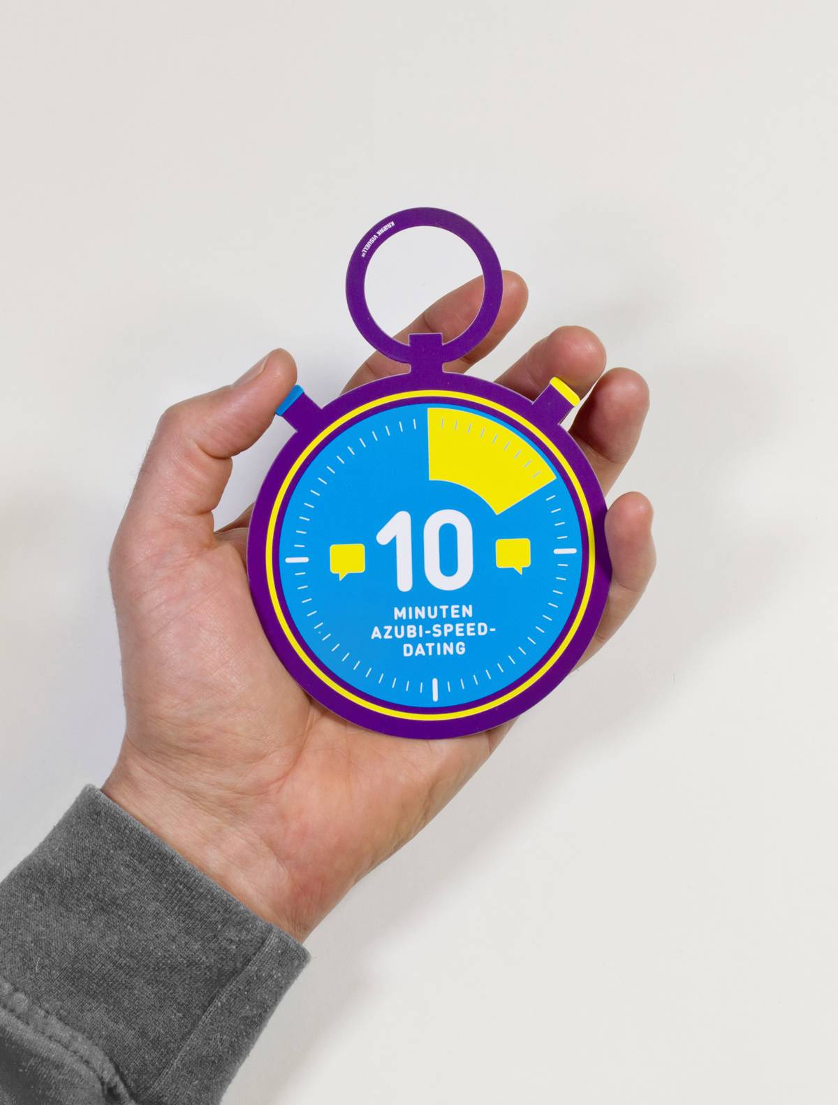 Eine Hand hält eine bunte Pappuhr mit der Aufschrift „10 Minuten Azubi-Speed-Dating“.