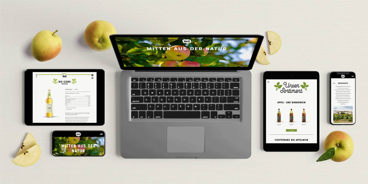 Laptop, zwei Tablets und zwei Smartphones liegen auf einem Tisch, alle zeigen eine Website mit Apfelwein-Produkten; ringsum liegen Äpfel und Apfelscheiben.