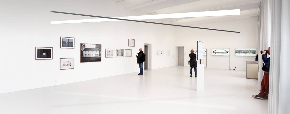 Heller Galerieraum mit weißen Wänden, an denen Kunstwerke hängen; mehrere Personen betrachten die Ausstellungsstücke, eine Person fotografiert.