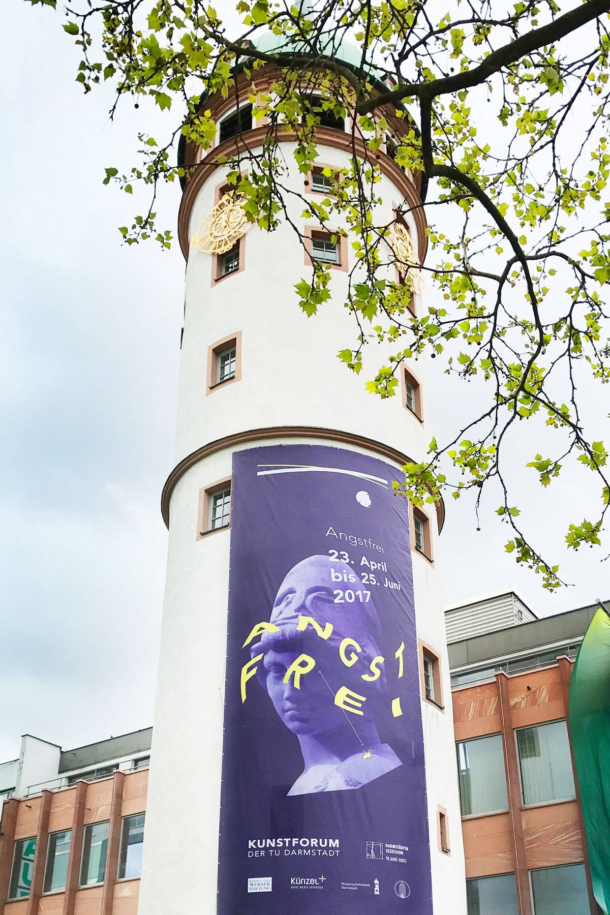 Weißer Turm mit violettem Banner für die Ausstellung „Angstfrei“ (23. April bis 25. Juni 2017) vor grünem Laub und grauem Himmel.