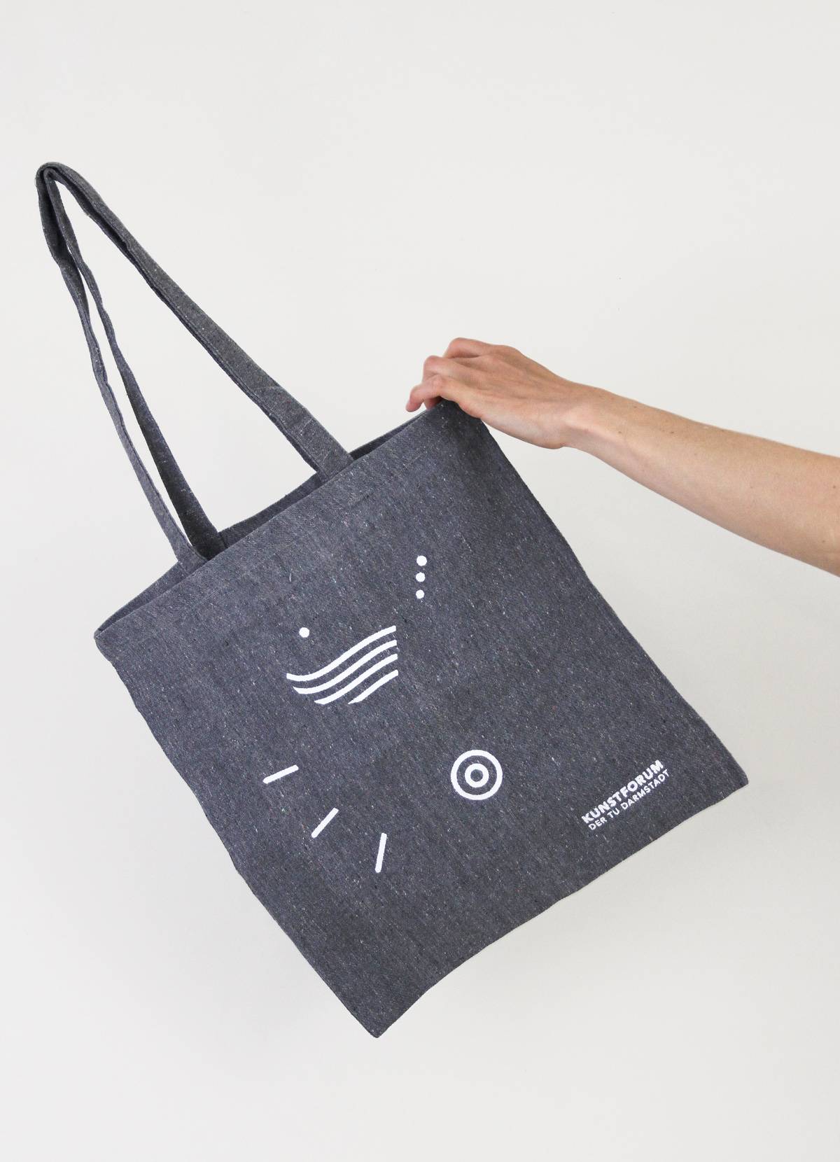 Eine Hand hält eine dunkelgraue Stofftasche mit weißen, abstrakten Symbolen und der Aufschrift „KUNSTFORUM DER TU DARMSTADT“.