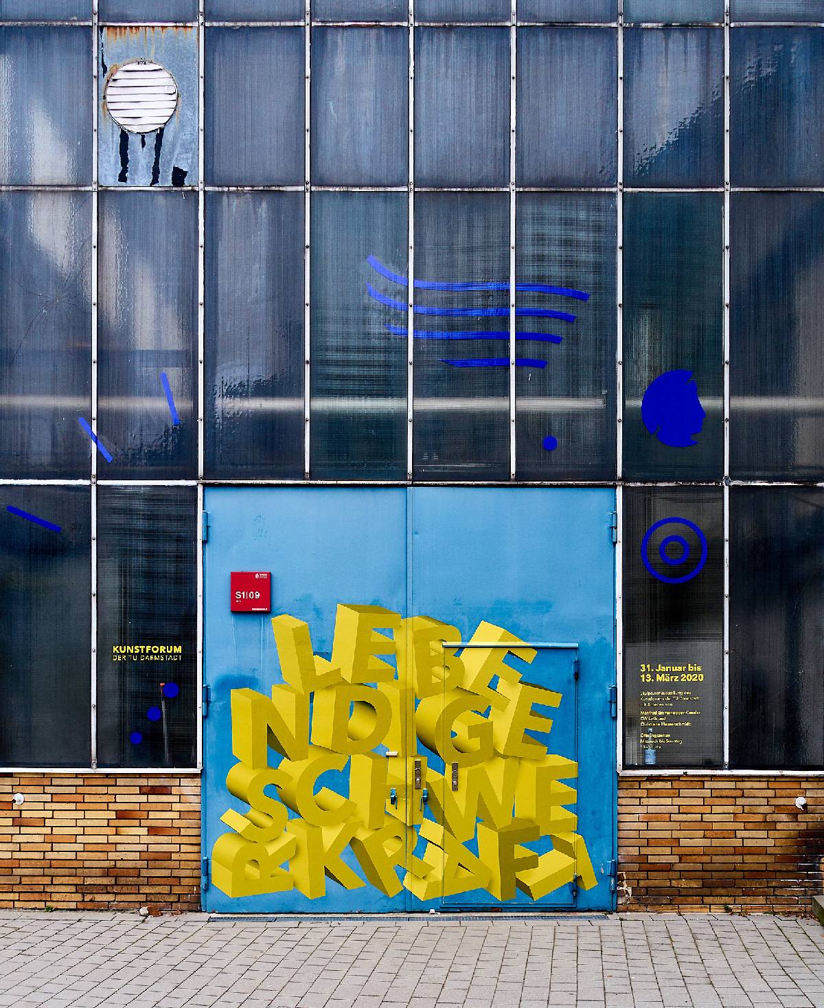 Glasfassade mit türkisblauer Tür, darauf gelbe 3D-Buchstaben „LEBENDIGE SCHWERKRAFT“, blaue grafische Elemente und Ausstellungsinformationen in Gelb und Weiß.
