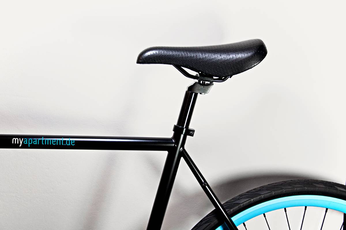 Detailaufnahme eines schwarzen Fahrrads mit schwarzem Sattel und hellblauem Reifen vor weißer Wand. Auf dem Rahmen steht "myapartment.de".