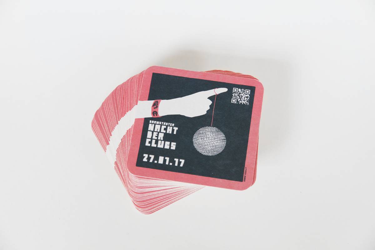 Ein Stapel Bierdeckel mit rotem Rand, darauf eine stilisierte Hand, die eine Diskokugel hält, daneben Text „Darmstädter Nacht der Clubs 27.01.17“ und ein QR-Code.