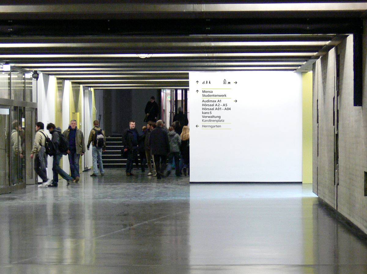 Menschen gehen durch einen modernen Flur mit Betonboden, an der Wand hängt ein großes weißes Wegweiser-Schild mit Orientierungspunkten wie Mensa, Hörsäle und Verwaltung.