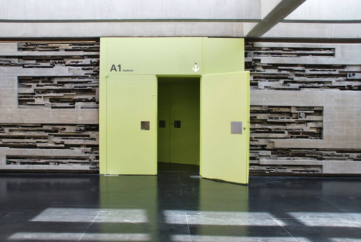 Große gelbe Doppeltür mit der Aufschrift „A1 Audimax“, teilweise geöffnet, vor einer Betonwand mit geometrischen Mustern.