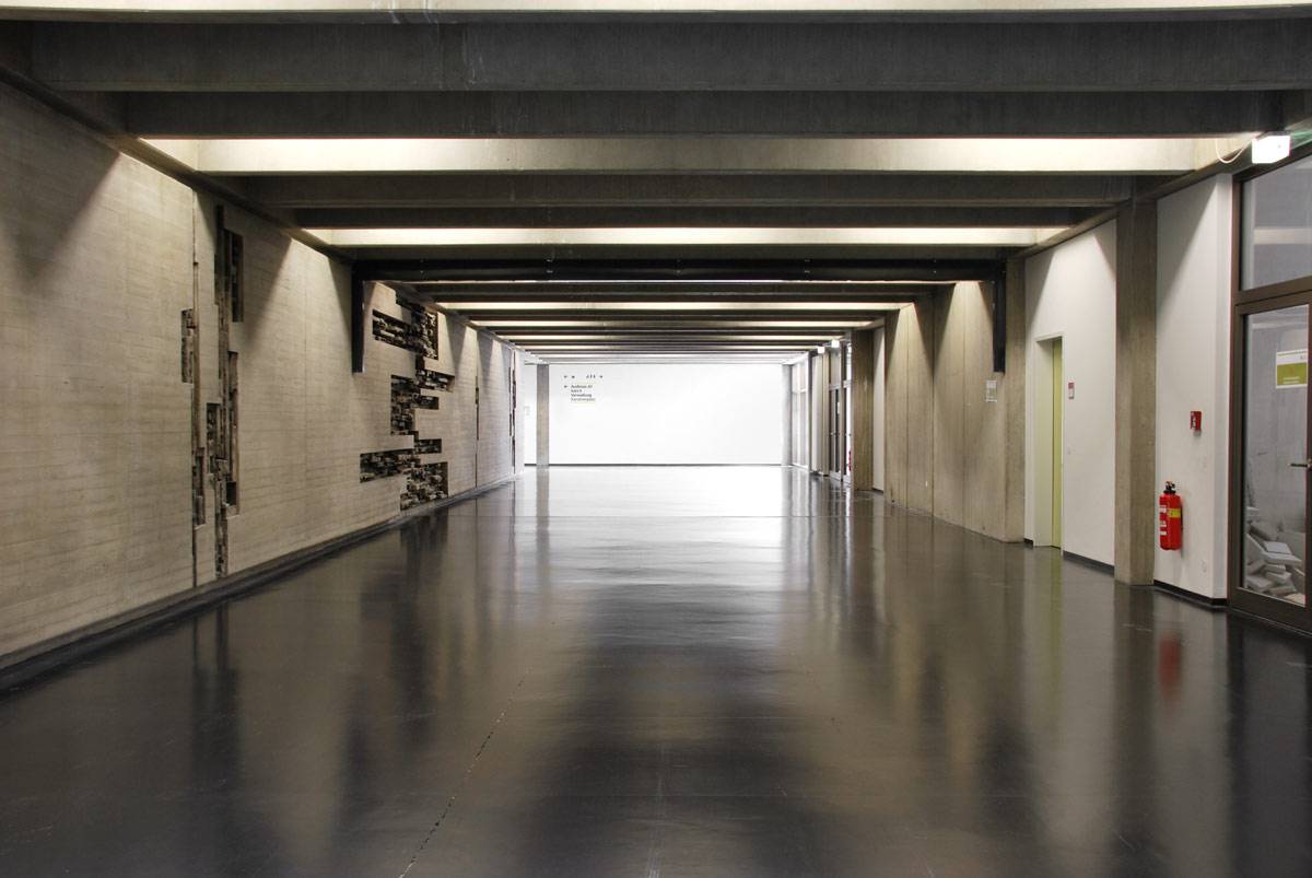 Langer, leerer Flur mit Betonwänden, dunklem glänzenden Boden und Deckenbalken; an einer Wand sind Kunstwerke, an der rechten Wand hängt ein Feuerlöscher.
