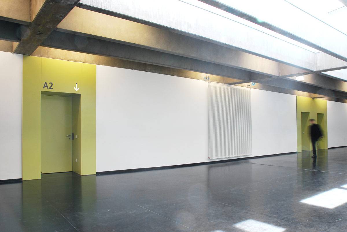 Heller Flur mit gelben Türen, einer mit der Beschriftung "A2", daneben eine weiße Wand und eine gehende Person im Hintergrund.