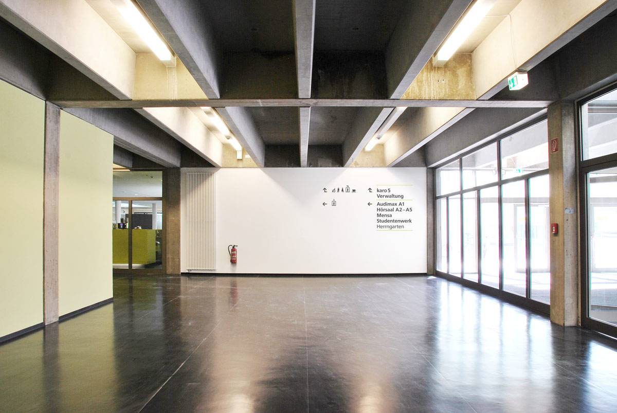 Heller, moderner Flur mit großen Fenstern, Sichtbetondecke und Wegweisern an einer weißen Wand.