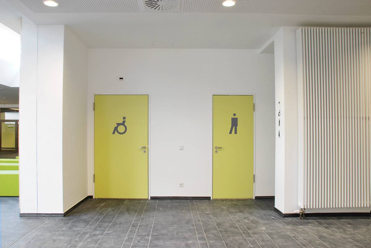 Zwei gelbe Türen in einem Flur, links mit Rollstuhlsymbol für barrierefreie Toilette, rechts mit Männersymbol für Herrentoilette.