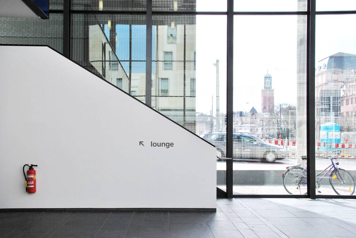 Heller Eingangsbereich mit weißer Wand, auf der ein Feuerlöscher und ein Schild mit Pfeil und dem Wort „lounge“ zu sehen sind; große Fenster geben den Blick auf eine Straße, ein Fahrrad und Gebäude frei.