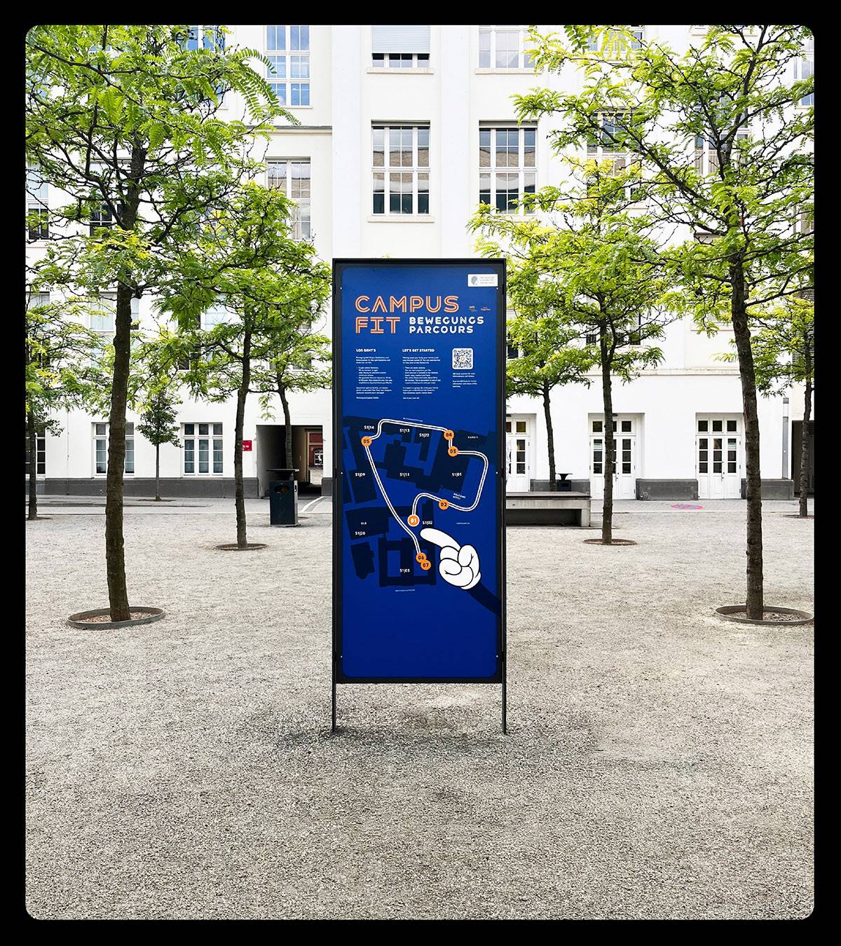 Infotafel mit der Aufschrift „Campus Fit Bewegungsparcours“ und einem Lageplan, aufgestellt auf einem Platz mit Bäumen vor einem weißen Gebäude.