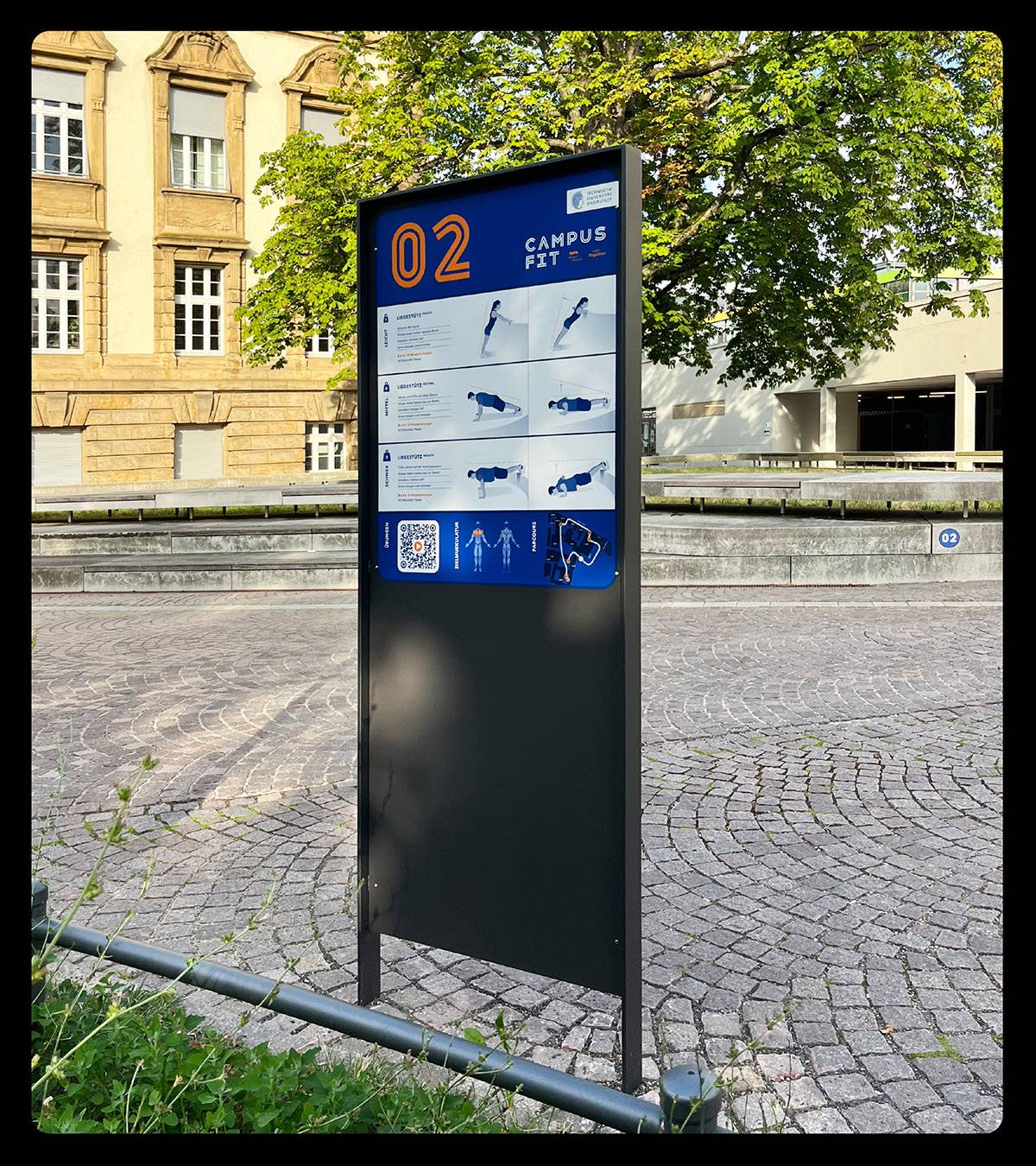 Metallschild mit der Aufschrift "02 Campus Fit" und bebilderten Anleitungen für Fitnessübungen, aufgestellt auf einem gepflasterten Platz vor einem Gebäude und Bäumen.