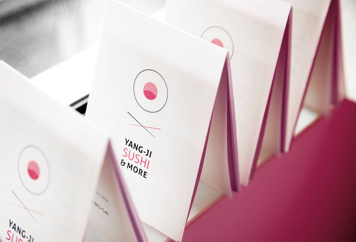Mehrere aufgestellte Speisekarten mit der Aufschrift "Yang-Ji Sushi & More" und minimalistischem Sushi-Logo in Rosa und Schwarz.