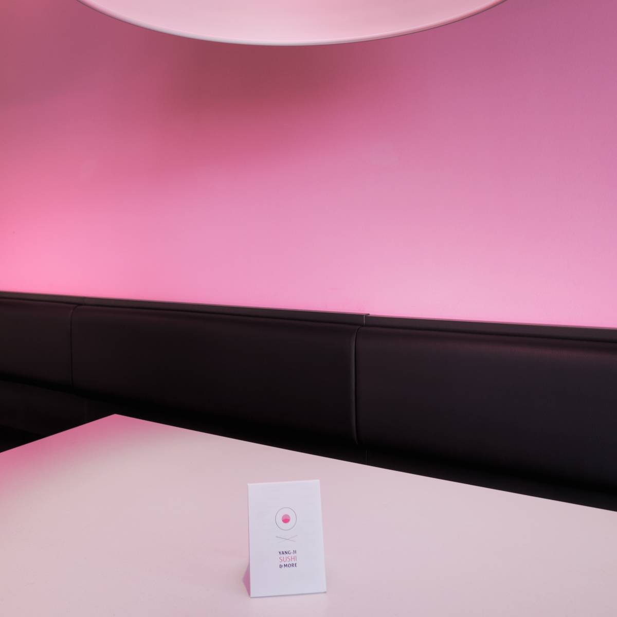 Weißer Tisch mit aufgestellter Speisekarte vor einer schwarzen Sitzbank, Hintergrund in rosa Licht getaucht.