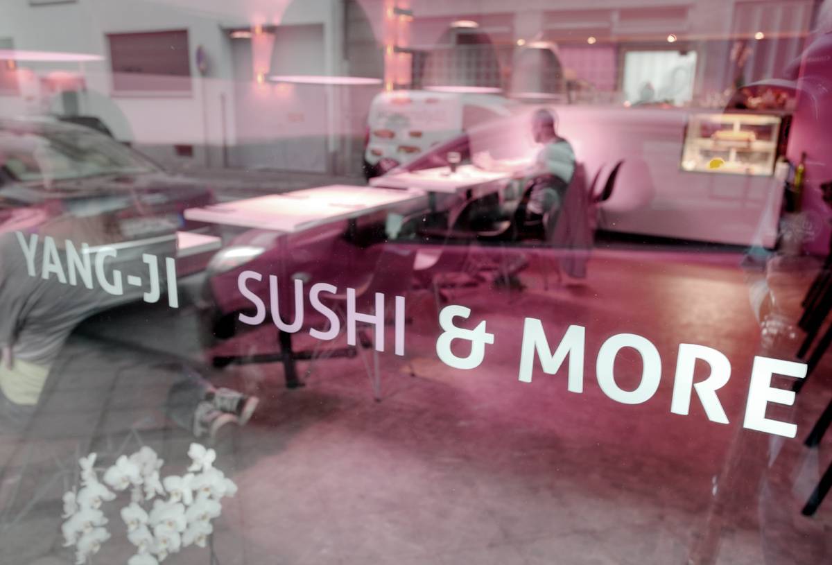 Schaufenster eines Sushi-Restaurants mit der Aufschrift „YANG-JI SUSHI & MORE“, im Hintergrund sitzt eine Person an einem Tisch.